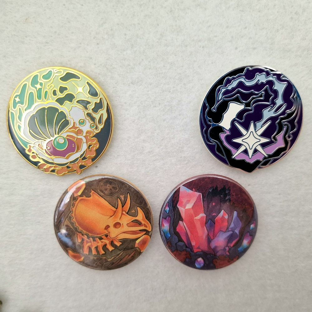 A photo of a pin board with the official Art Fight Seafoam and Stardust enamel pins, with unofficial Fossils and Crystals badge pins underneath.