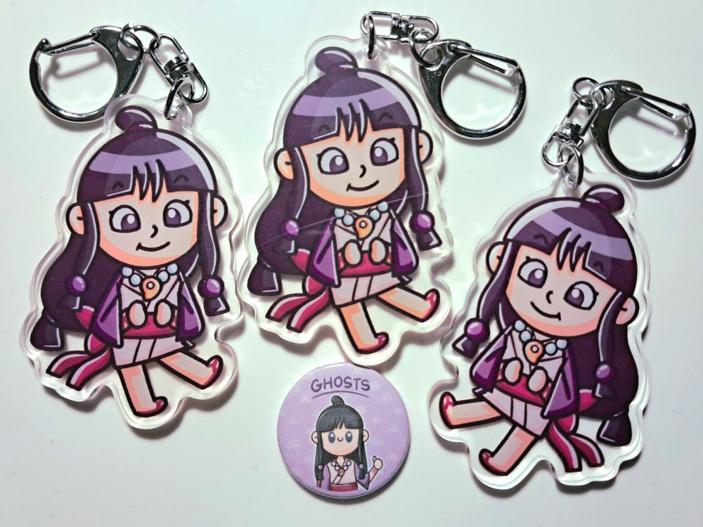 A photo of three acrylic keyrings and one small badge from my Etsy shop, all of Maya Fey from Ace Attorney. The badge has the word GHOSTS above Maya's head, and she's doing a thumbs up.