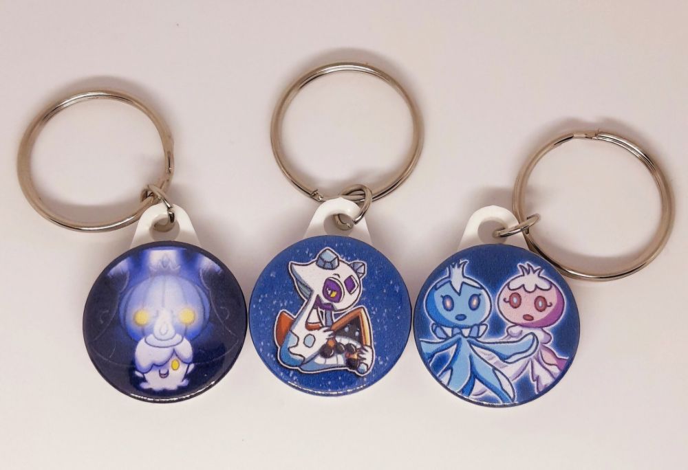 A photo of three Pokémon badge keyrings from my Etsy shop. The first is of Litwick and Chandelure, the second is of Frosslass holding a Snorunt, and the third is of a pair of Frillish - one blue male and one pink female.