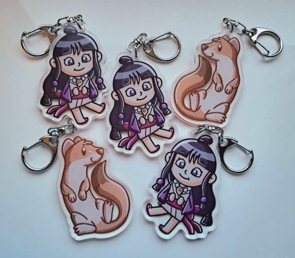 A photo of five acrylic keychains from my Etsy shop. Two of them are a cute stoat, and three of them are Maya Fey from Ace Attorney.