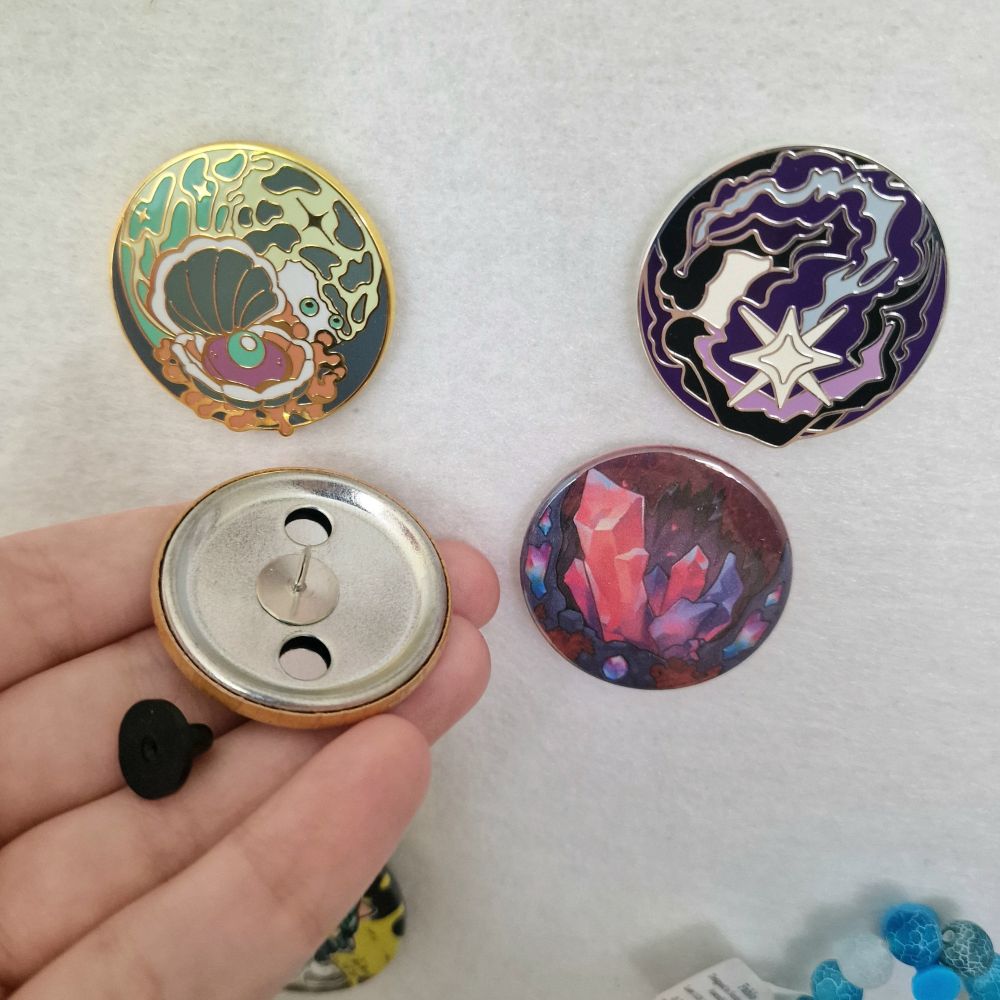 A photo of a pin board with the official Art Fight Seafoam and Stardust enamel pins, with unofficial Fossils and Crystals badge pins underneath. The Fossils pin has been pulled off in this one to reveal a badge back with a pin glued to it, and a black rubber back to stick on the pin.