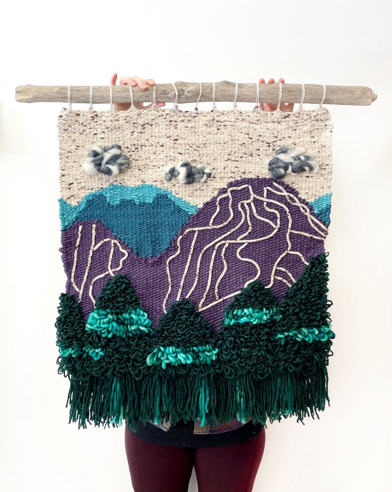 A person holds a weaving in front of them. It hangs from a piece of great driftwood. The weaving begins at the base with fluffy green triangles for trees, then a series of mountains in purple & blue, then a cream sky with clouds. On the foreground purple mountain there are cream lines.