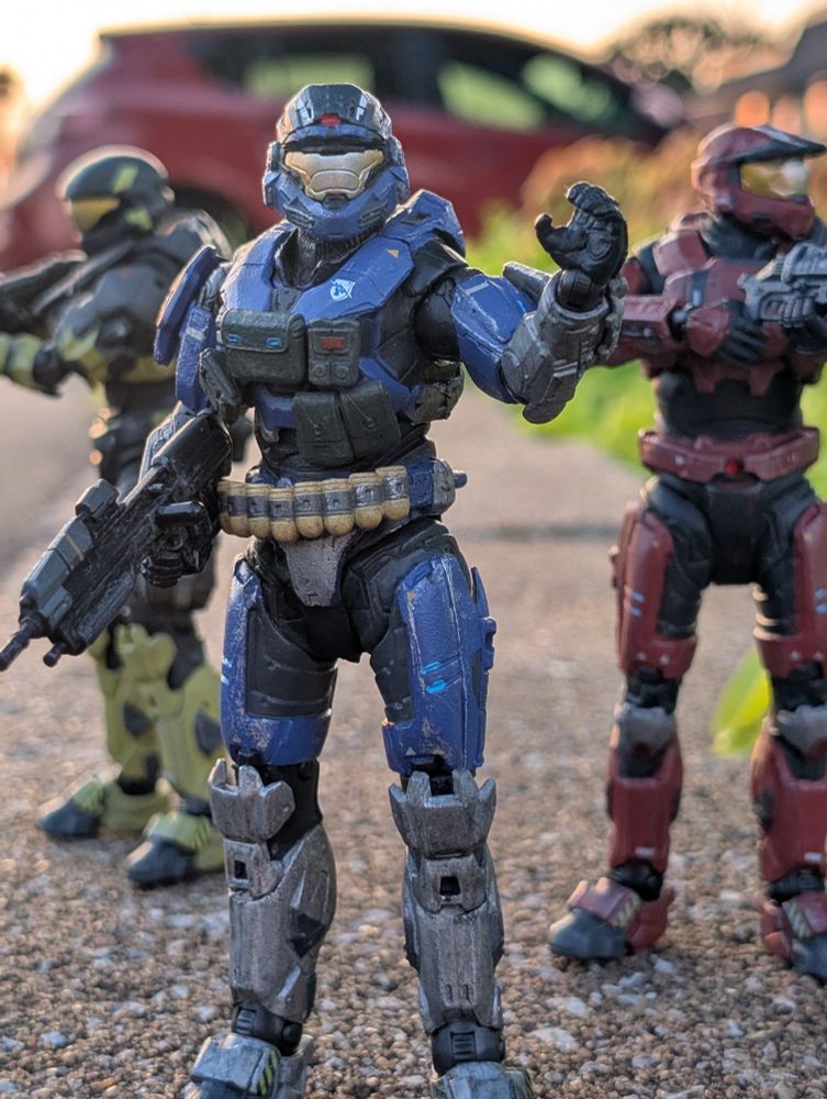 Three Halo Spartans backlit by sunset. The center one holds his curled hand up as if to signal the two behind him, which are holding defensive stances in either direction towards the edge of the frame. 