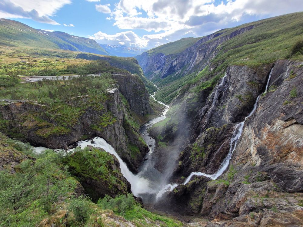 måbødalen, western norway. on the left is vøringsfossen, a popular tourist attraction that would have looked a lot better if the river weren't dammed. it's still a ridiculously picturesque location where two waterfalls meet at the bottom of a steep-walled valley and wind towards the horizon. in the far background are mountains with some remaining patches of snow, shaded blue by haze and distance giving a postcard-y feel. above is a mix of blue sky and clouds partly illuminated by the sun