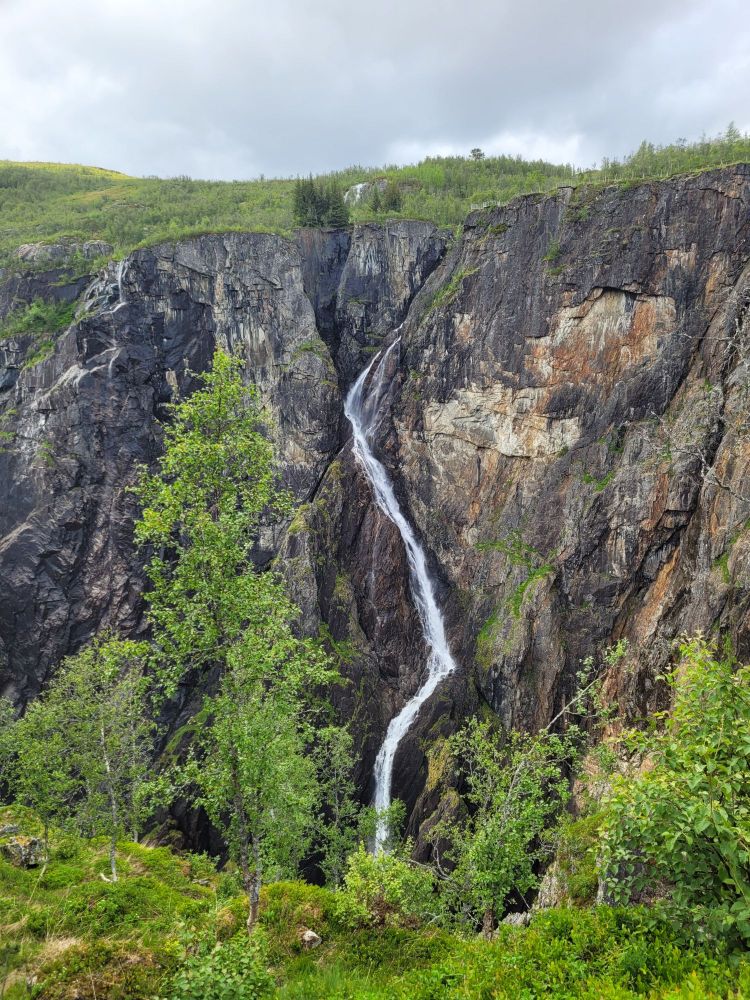 a small waterfall zig-zagging its way down a near-vertical cliff. this is the waterfall on the right in the next picture. the cliff face is sandwiched in between foreground foliage and background foliage beneath cloudy skies