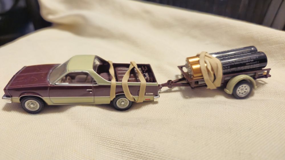 A Matchbox car El Camino, brown with tan top, towing a trailer that carries two AA batteries rubber-banded onto the trailer. It's an electric El Camino (sort of)!