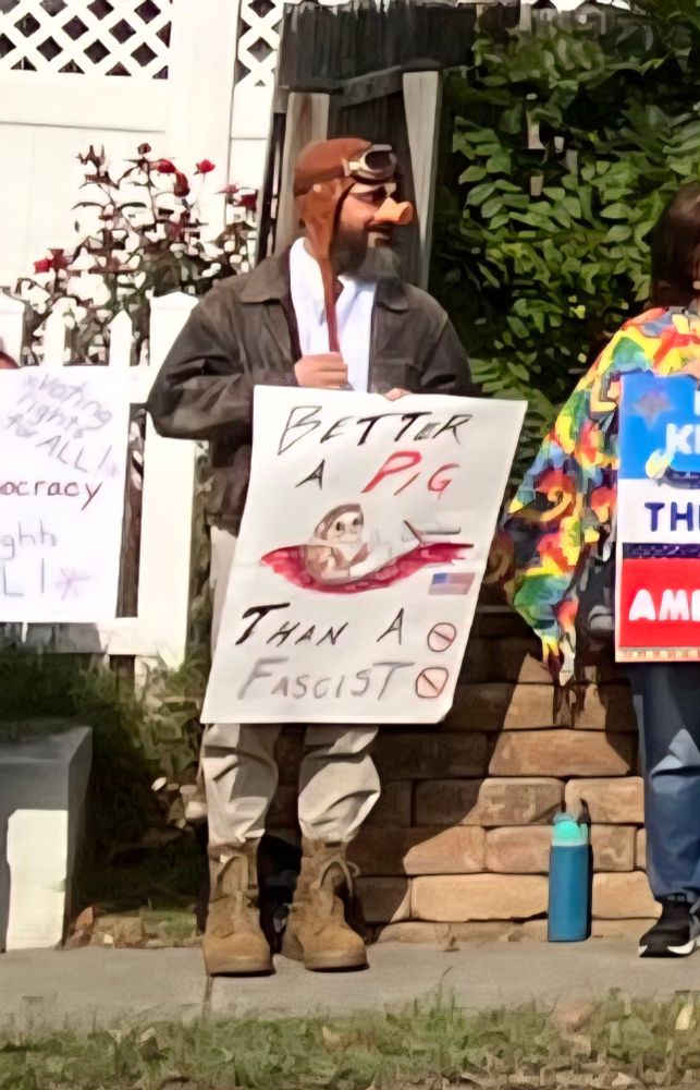 A man standing on a sidewalk, between two other people who are cut off. He is wearing an old-timey aviator cap, brown leather with straps hanging down and goggles pushed up onto the forehead. He has a pig nose and a beard. The rest of his outfit is mostly covered by his sign, but it includes a worn leather bomber jacket over a white shirt, tan pants, and brown lace-up boots. His sign is hand-written and hand-drawn. It reads: "Better a pig than a fascist." In the middle of the sign is a drawing of a pig in the cockpit of an old plane. The pig is dressed just like the man is. There is a small American flag below the drawing. It refers to the Studio Ghibli film "Porco Rosso."