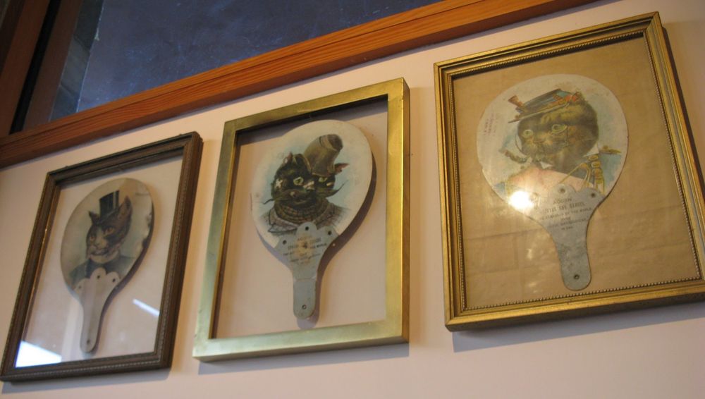 Three rectangular cases with gold edges, in portrait orientation. Each has a fan shaped like a ping-pong paddle. Each fan has a cat in a hat, but not like Dr. Seuss. These cats look Victorian. The first is a gentleman. He is wearing a small black top hat between his ears and a black top with a white collar and a white front. His head is brown except for the area between his eyes and around his mouth, which is white. 
The second cat has been through it. His hat is floppy and askew. His ears are ragged. He has little bandages on his nose and cheeks, and his whiskers are twirled into a long, wonky mustache. He wears a red-and-green plaid jacket buttoned up to his neck with a plaid scarf underneath. 
The third cat seems to be a soldier. He wears a flat blue cap with red trim and a blue jacket with gold epaulets and a red sash. It's hard to see much detail because the light is reflecting off the glass.