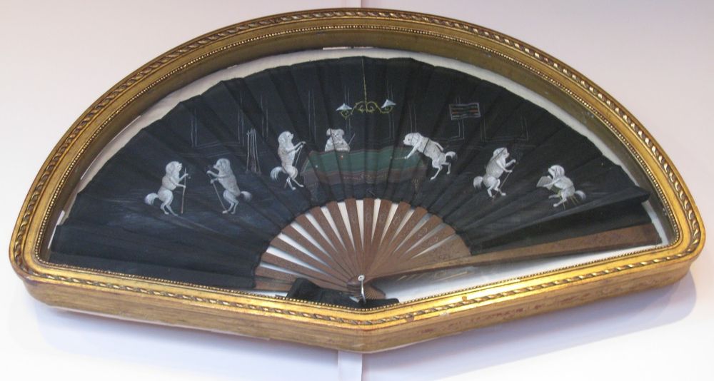 A hand fan in a hand fan-shaped gold-edged case. The fan is spread out. It has a black background and brown wood slats. On the fan, white dogs, all standing on their hind legs, are playing pool. Three dogs are at the pool table -- one is lining up his shot while the other two watch. To the left, two dogs face each other while chalking their pool cues. To the right of the pool table, another dog is chalking its cue while the last dog sits on a stool with a piece of paper, maybe keeping score.