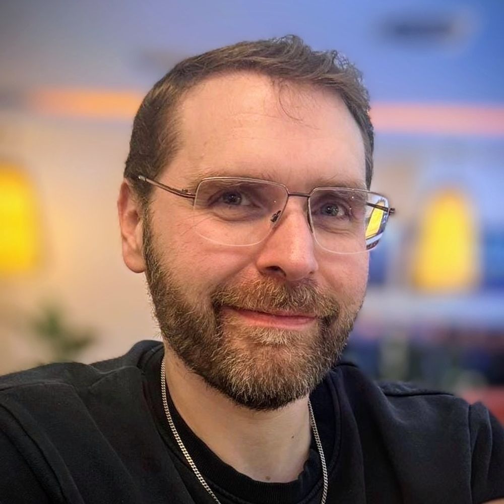 Porträt eines Mannes mit Bart und Brille, der entspannt in die Kamera lächelt. Der Hintergrund ist leicht verschwommen, warmes Licht verleiht dem Bild eine gemütliche Atmosphäre.