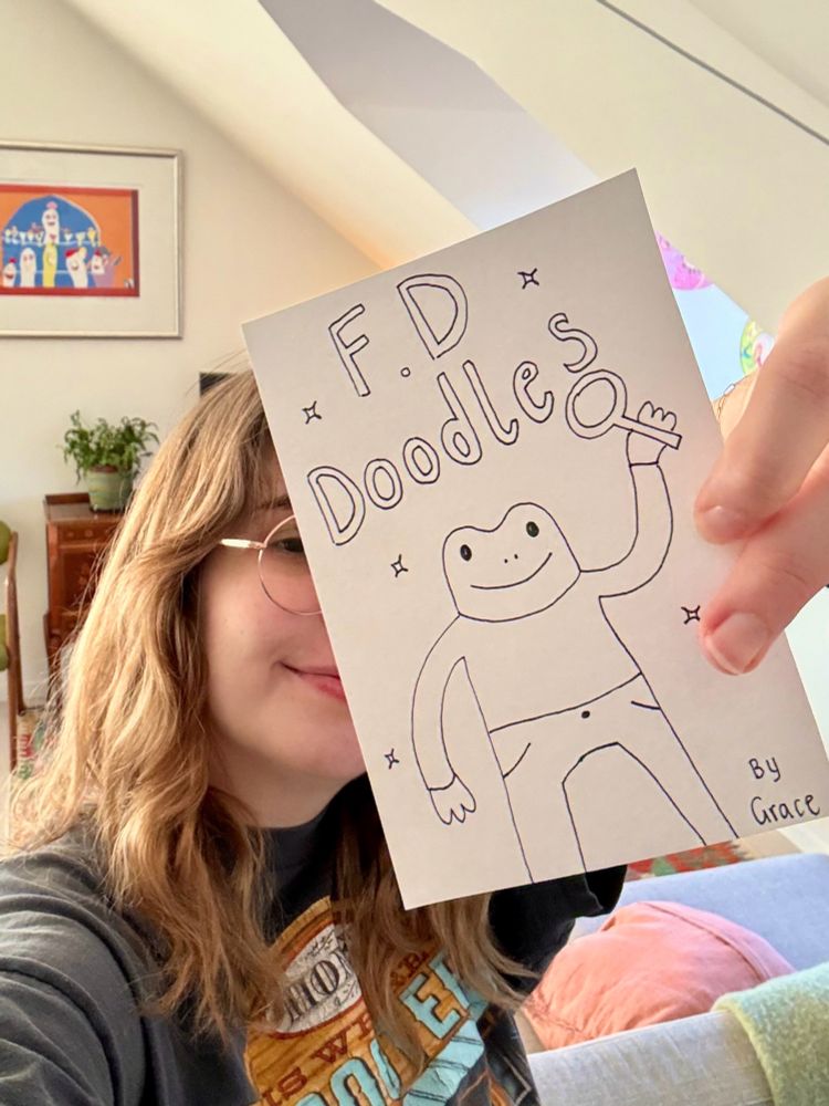 grace holding a small piece of paper with a drawing of frog detective that says “F.D Doodles”