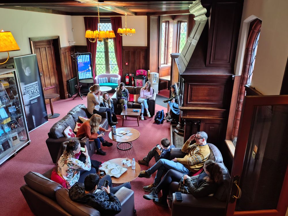 Open hall of Foxhill House with students relaxing on leather sofas in front of a large fireplace with ornate wood surround. 