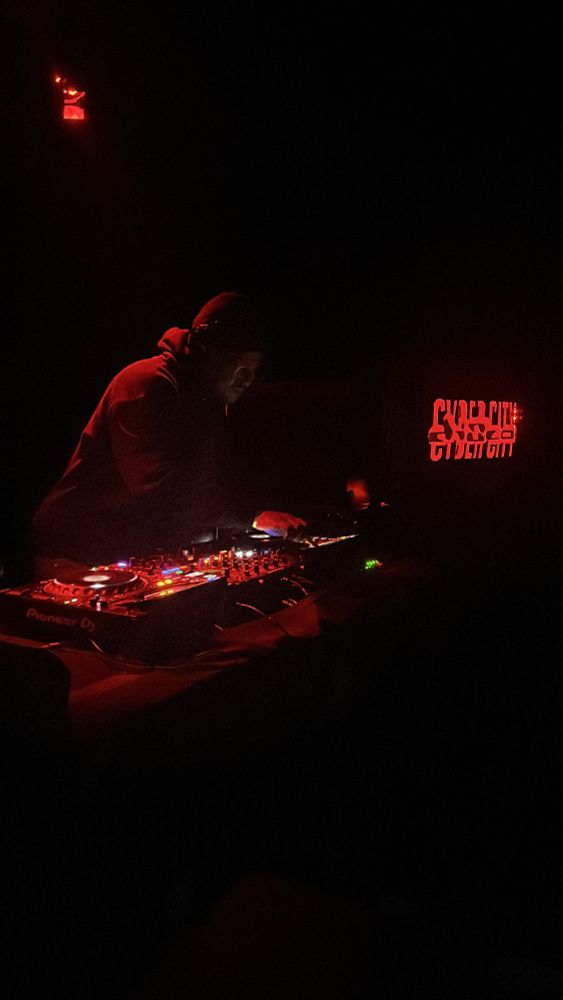 DJ Ariesfirebomb under a red light mixing dance music at Cyber City Disco. A red neon sign to their right reads Cyber City Disco