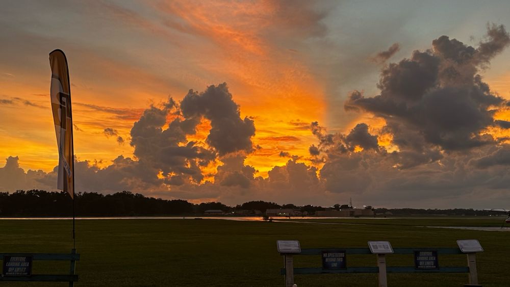 Striking orange sunset behind puffy clouds in the distance. Closer to the camera, the sky is clear and transitions to shades of blue. Skydive City (Z-hills) landing area is in the foreground.