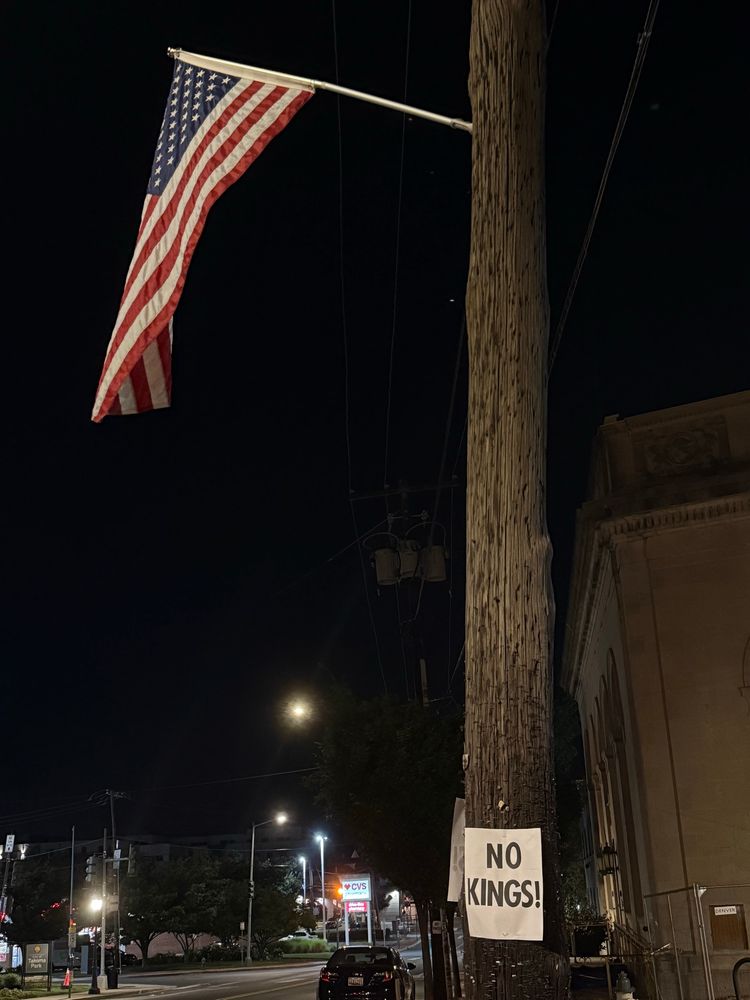 Picture of a lightpole at night on a city street. At the top of the pole is an American flag waving in the breeze. And the bottom of the pole is a sign printed on computer paper that reads “NO KINGS!”