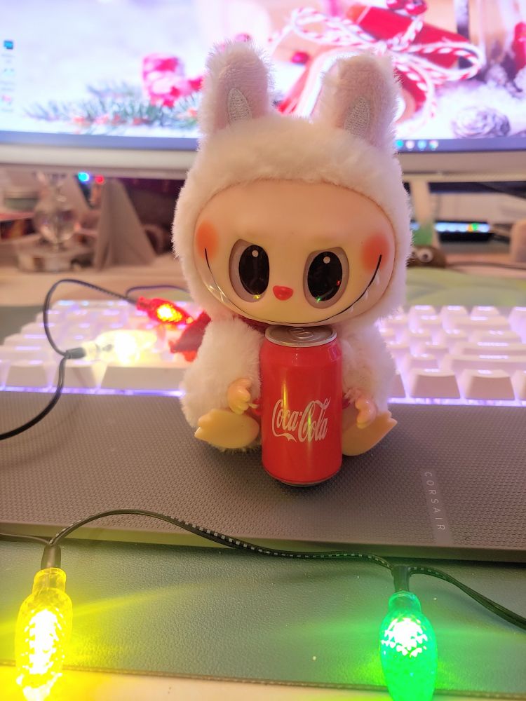 Photo (by me) of a Labubu doll holding a can of Coca-Cola, sitting on a keyboard surrounded by multi color Christmas lights.