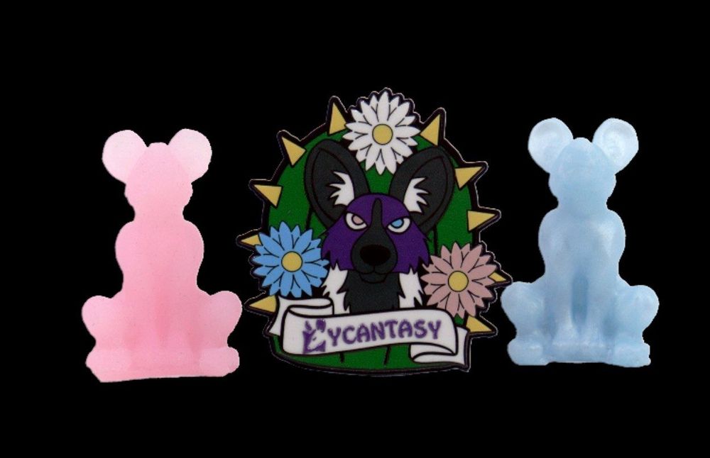 An enamel pin of a purple African wild dog in front of a cactus with pink white and blue blossoms and a banner that says Lycantasy in the foreground with two Mini silicone Lyca's next to it
