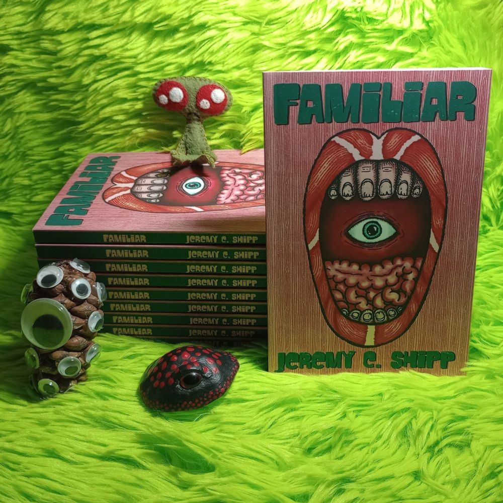 Here's a photo featuring a stack of the book Familiar by Jeremy C. Shipp. There's another copy leaning against the stack, with the cover facing the camera. The cover shows an open mouth with lips made of muscle and teeth made of fingers and a tongue made of intestines. In the back of the mouth, there's an eye. 

My fabric art homunculus is sitting on the stack of books. She's green with big sparkling red eyes and a tiny frown. There's also a black and red mushroom near the stack with a large eye. Near the stack, there's also a pinecone covered with googly eyes.