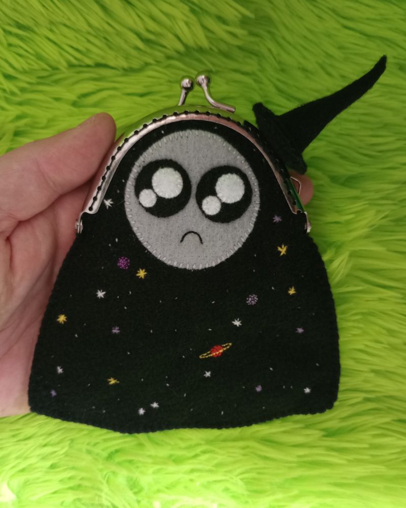 Here's a lovely photo of the cosmic ghost bag. She's a black ghost with various stars and planets embroidered on her body. There are little white stars, larger white stars, yellow stars, purple stars, a dark purple planet, a light purple planet, a red planet with a yellow ring. She has a gray moon-like face with large sparkly black eyes and a little frown. She's wearing a black fabric witch hat. She's resting on my palm and there's green fur in the background of the pic.