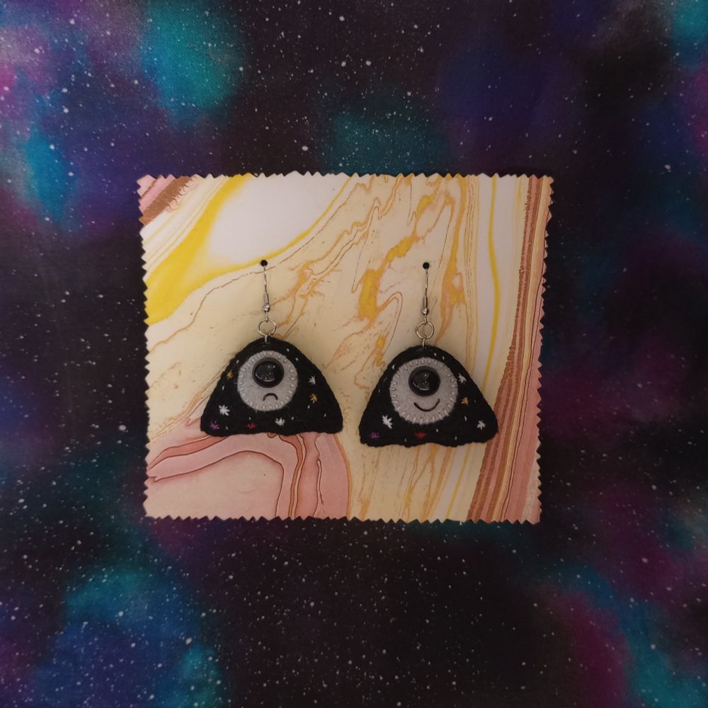 Here's a lovely photo of my fabric art cosmic ghost earrings. They're little black ghosts with gray moon faces. Their bodies are full of embroidered white, purple, red, and yellow stars. They each have a bulbous black eye. One has a little frown and one has a little smile. In the background of the pic is a galaxy print fabric.