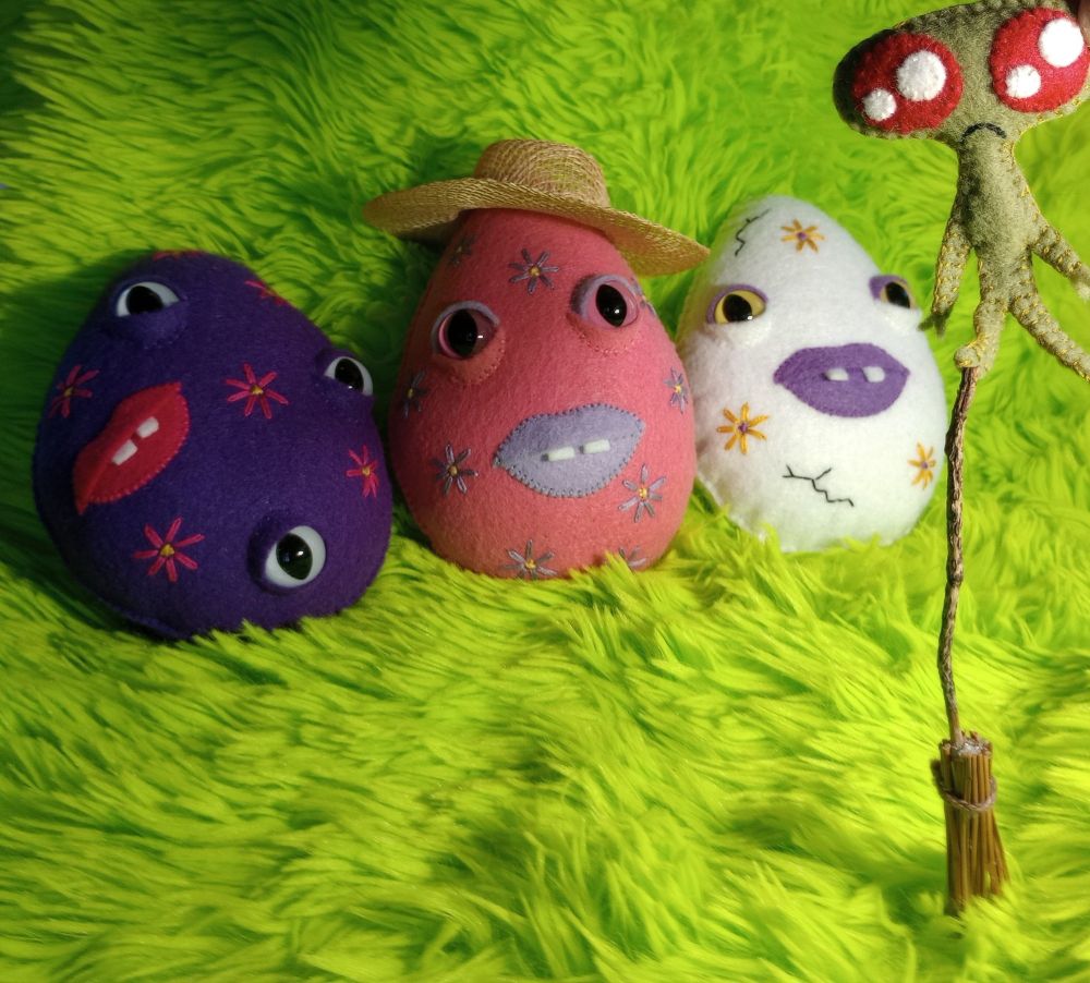 Here's a lovely photo of three fabric art eggs watching the tiny witch homunculus balancing on one foot on a small broom. The eggs are purple, pink and white, and they're covered with embroidered flowers. The eggs have mouths with lip colors that match their flowers. They have two flat teeth poking out of their mouths. The pink egg is wearing a small sun hat. The tiny witch homunculus is green with large sparkly red eyes and a little frown. There's green fur in the background of the photo.