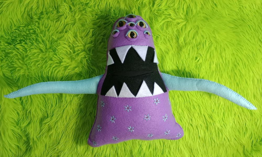 Here's a lovely photo of Big Bad Beatrice. She's a tall, purple ghost-like being, with seven yellow cat-like eyes and greenish blue eyeshadow. She has a large mouth filled with big, sharp teeth. She has two greenish blue tentacle-like arms. Her lower body is covered with embroidered greenish blue flowers. There's green fur in the background of the photo.