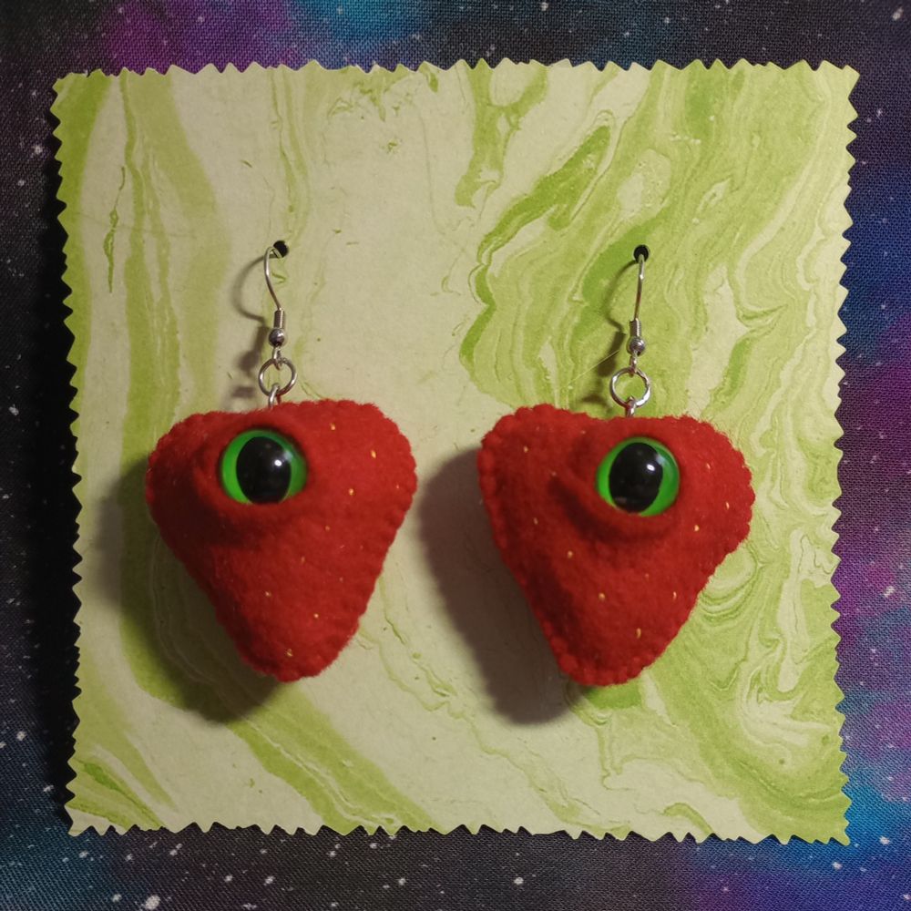 Here's a lovely photo of my fabric art strawberry earrings. They're red strawberries with hand-embroidered yellow seeds and bulbous green and black eyes. They're dangling on a swirly green card with spiky edges.