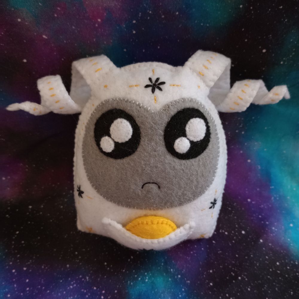 Here's a lovely photo of Ambrosia, my fabric art phantom. She's a white ghost with a gray face, large sparkly black eyes, a little frown, and a pouch over her belly. There's a golden fabric orb in her pouch. Ambrosia is covered with hand-embroidered mystical symbols. There are black flowers with golden lines around them. She has two twirly wisps coming out of her head. There's a lovely galaxy print fabric in the background.