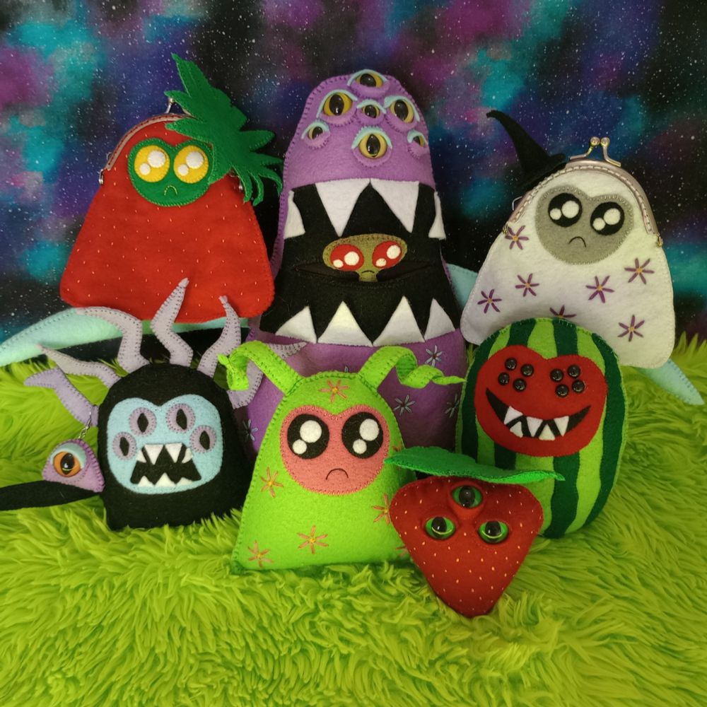 Here's a lovely photo of a plethora of hand-sewn fabric art creatures. There's a watermelon creature with a red face, eight black seed-like eyes, and a wide mouth with five sharp teeth. There's a white ghost trinket bag covered with purple embroidered flowers. She has a gray face, sparkly black eyes and a little frown. There's a strawberry creature with three bulbous green eyes. There's a watermelon ghost, and a black creature with sharp teeth wearing a bulbous eye earring. There's a large purple creature with many yellow eyes and an open mouth, with a little green creature popping her head out. All the creatures are sitting on green fur and there's a galaxy print fabric in the background.
