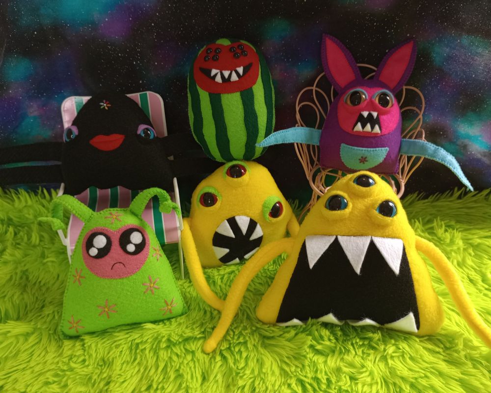 Here's a lovely photo of some of my fabric art creatures. They're on green fur with a beautiful fabric galaxy in the background. There's a fabric art watermelon with eight eyes and a wide toothy grin. There's a black phantom with purple eyeshadow, red lips, and four tentacle-like arms. She has an embroidered flower on her forehead where each petal is a different color of the rainbow m there's a watermelon ghost, two yellow blob like creatures. They each have three eyes, two tentacles, and open mouths full of sharp teeth. There's also a rabbit-like creature with a purple body, a pink face, and sharp tooth. The rabbit also has two blue tentacles and a blue pouch over the belly with an embroidered pink flower on it.