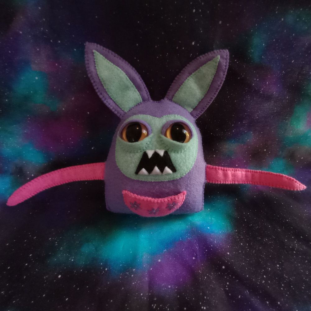Here's a lovely photo of Sweet Theodore. He's a purple rabbit with a greenish blue face, large dark yellow eyes, and an open mouth filled with sharp teeth. He has two pink tentacles and a pink pouch over his belly with three purple embroidered flowers. There's a galaxy print fabric in the background of the photo.