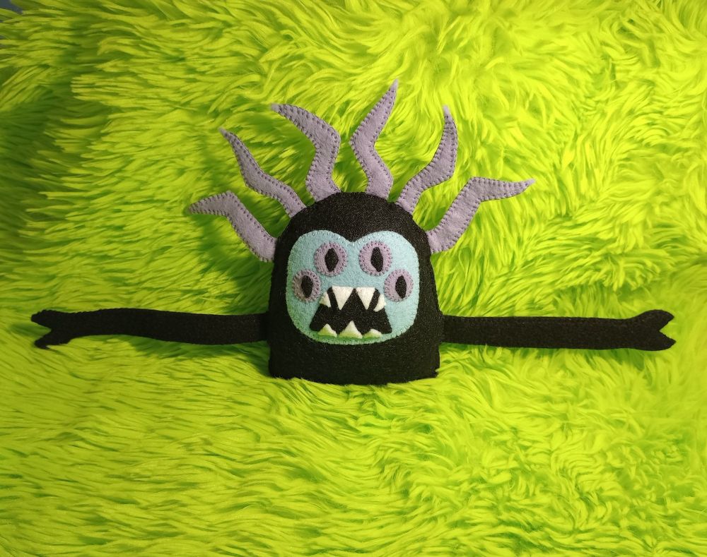 Here's a lovely photo of my fabric art creature Sweet William. He has a black gumdrop-shaped body and a blue face with four lavender eyes. He has an open mouth and seven sharp teeth. He has long arms with hands that are like pincers. He has six lavender, lightning-shaped spikes coming out of the top of his head. There's green fur in the background of the photo.