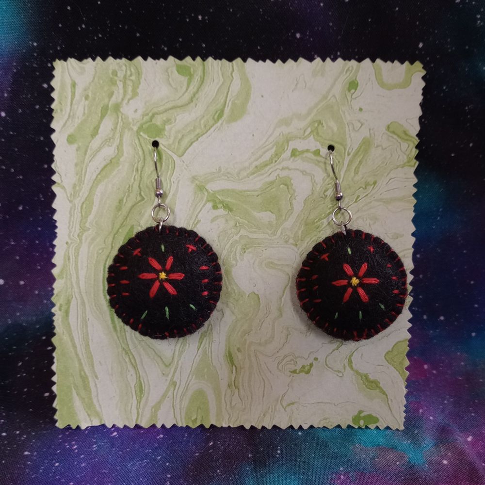 Here's a lovely photo of my fabric art earrings featuring a red flower and a yellow center. The fabric earrings are black circles with a flower in the center, surrounded by embroidered lines of red and green. The earrings are displayed on swirly green marble paper. There's a beautiful galaxy print fabric in the background.