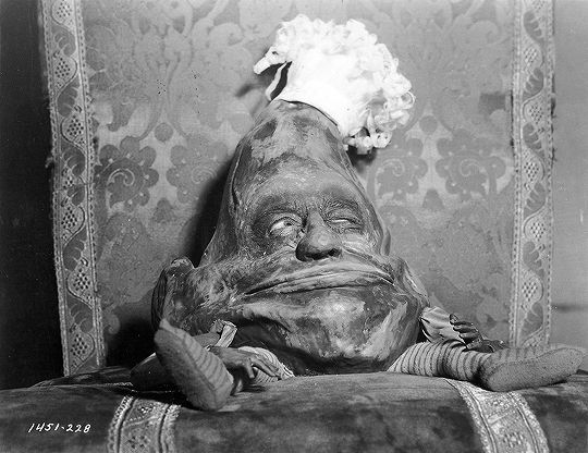Here's a lovely black and white pic from the old live action Alice in Wonderland film. It features an anthropomorphic pudding or something of the sort. He has gangly arms and legs. He's sitting on a chair too large for his body.