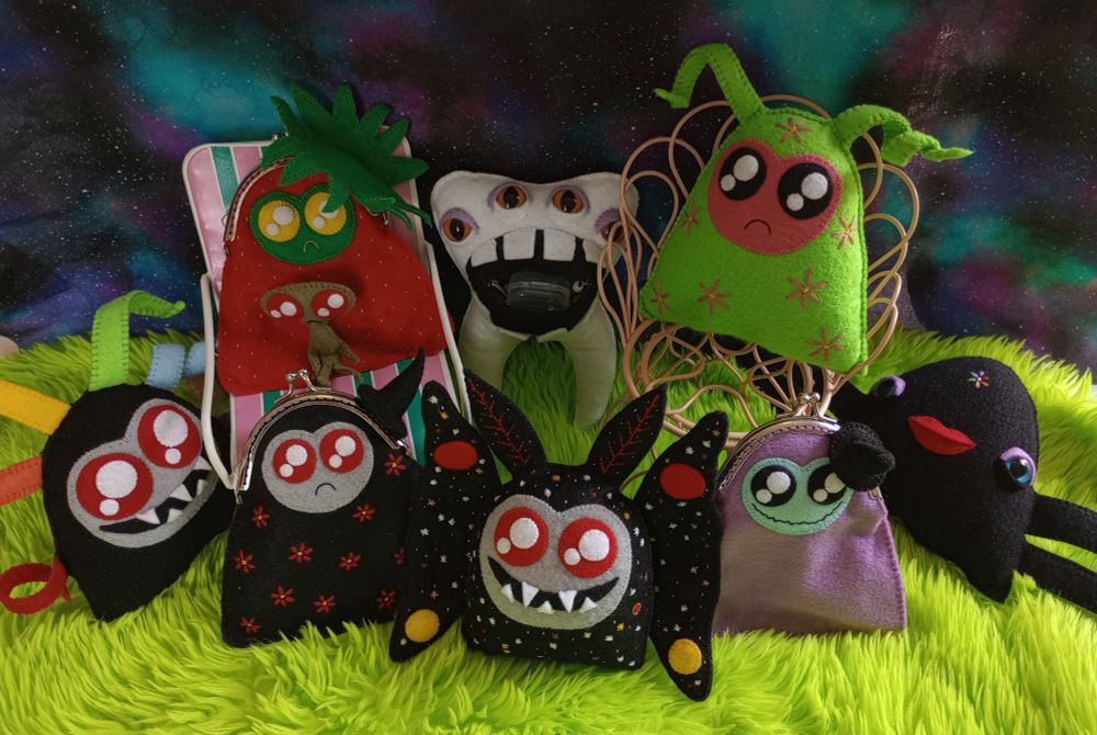 Here's a lovely photo of a plethora of my fabric art creatures. They're on green fur with a galaxy print fabric in the background. There's a black cosmic moth with a gray face, twinkly red eyes, and a wide smile full of sharp teeth. She's covered with embroidered stars and planets of various sizes and colors. There's a watermelon ghost covered with embroidered pink flowers sitting on a pink wire chair. There's a watermelon ghost bag and a small homunculus sitting on a pink and green striped chair. There's a tooth creature with a wide open mouth and four orange eyes and lavender eyeshadow. There's a black phantom with a gray face and wisps of every color of the rainbow.