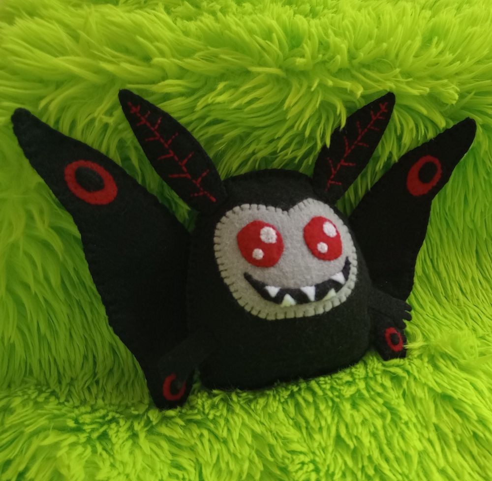 Here's a lovely photo of my fabric art Mothman. He's a black Mothman-like creature with a gray face, twinkly red eyes, and a wide grin full of sharp teeth. His wings have red and black circular markings on the tips.