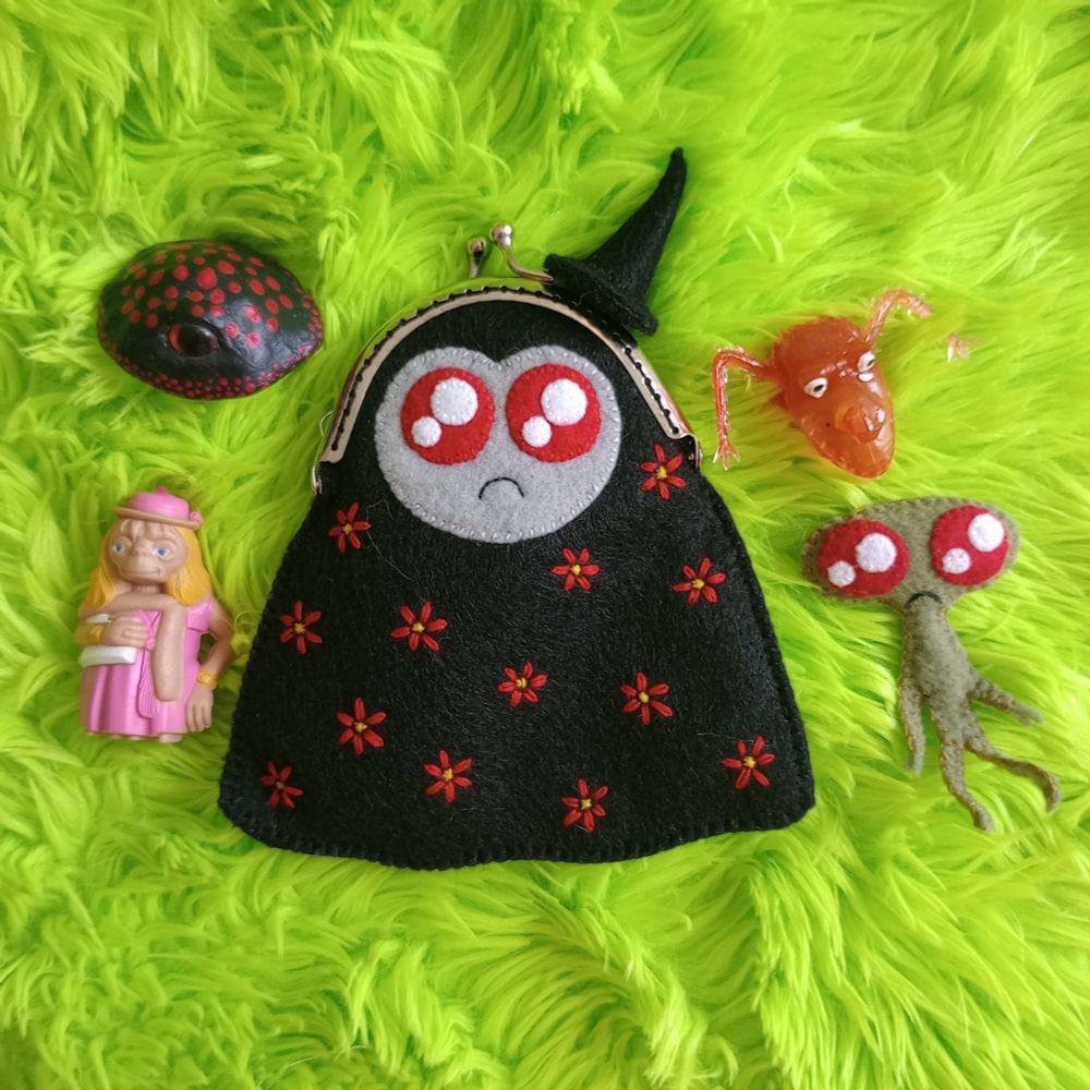 Here's a lovely photo of the fabric art ghost bag. She's a black ghost with a gray face, twinkly red eyes, and a little frown. The top of her head is a silver-colored coin purse clasp with a fabric, magnetic witch hat attached to it. She's covered with embroidered red flowers. There's green fur in the background of the pic. All around her are the items I mentioned in the skeet: a fabric art green homunculus with twinkly red eyes and a little frown, a red vintage monster finger puppets, a black mushroom sculpture with one eye, and a little ET figurine where he's dressed up as a lady.