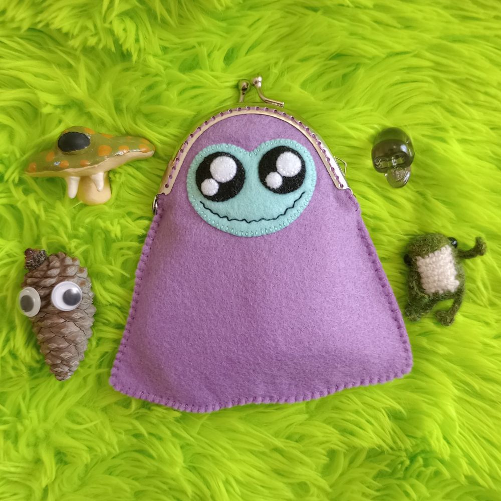 Here's a lovely photo of Madeline. She's a purple fabric art ghost bag, with a greenish blue face and large sparkly black eyes. She has a wide zigzagging sort of grin. The top of her is a silver-colored metal coin purse clasp. There's green fur in the background of the pic. Around her are a green and yellow mushroom sculpture with one back eye, a pinecone with two googly eyes, a transparent skull, and a little knitted frog.