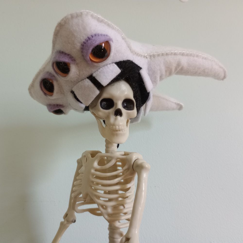 Here's a lovely photo of Tooth gnawing on the head of a small plastic skeleton.There's a light blue wall in the background. Tooth is a fabric art tooth with four orange eyes and lavender eyeshadow. He has a big mouth with thick molars.