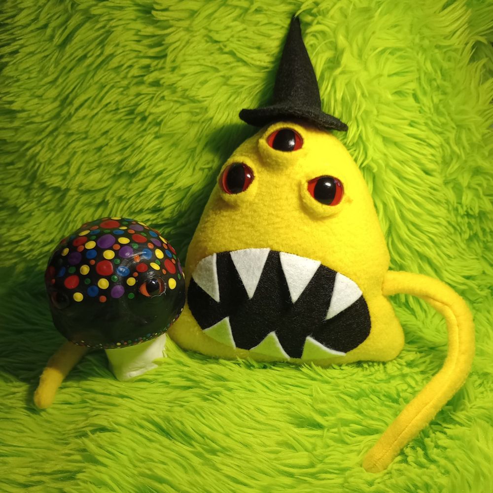 Here's a lovely photo of Lemondrop and Max. Lemondrop is a fabric art creature with three bulbous red eyes, a big oval mouth full of sharp teeth. He has two thin tentacle-like arms. He's wearing a black fabric witch hat that I hand-sewed. He has an arm around his buddy Max. Max is a clay mushroom creature that I sculpted with two eyes. He's a black mushroom with rainbow dots painted on the top of his head 