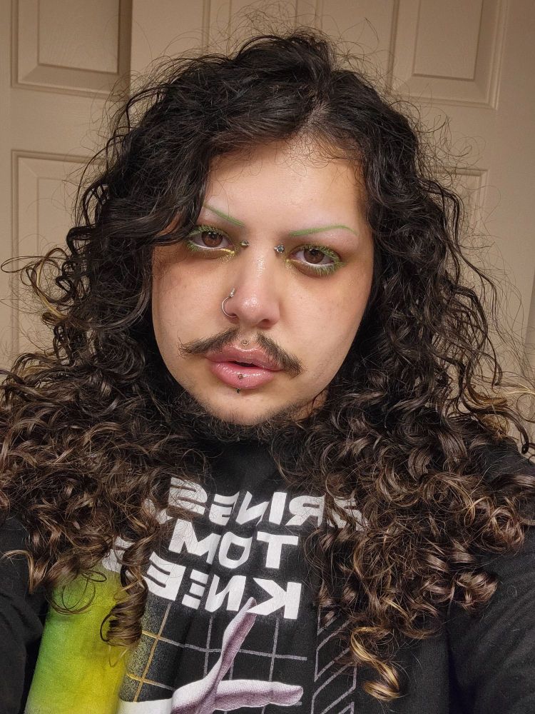 Makeup featuring a thin green brow with matching eyelashes, white eyeliner in the waterline, orange blush and silver lipgloss with shimmery green eyeshadow and grungey green tones in the crease