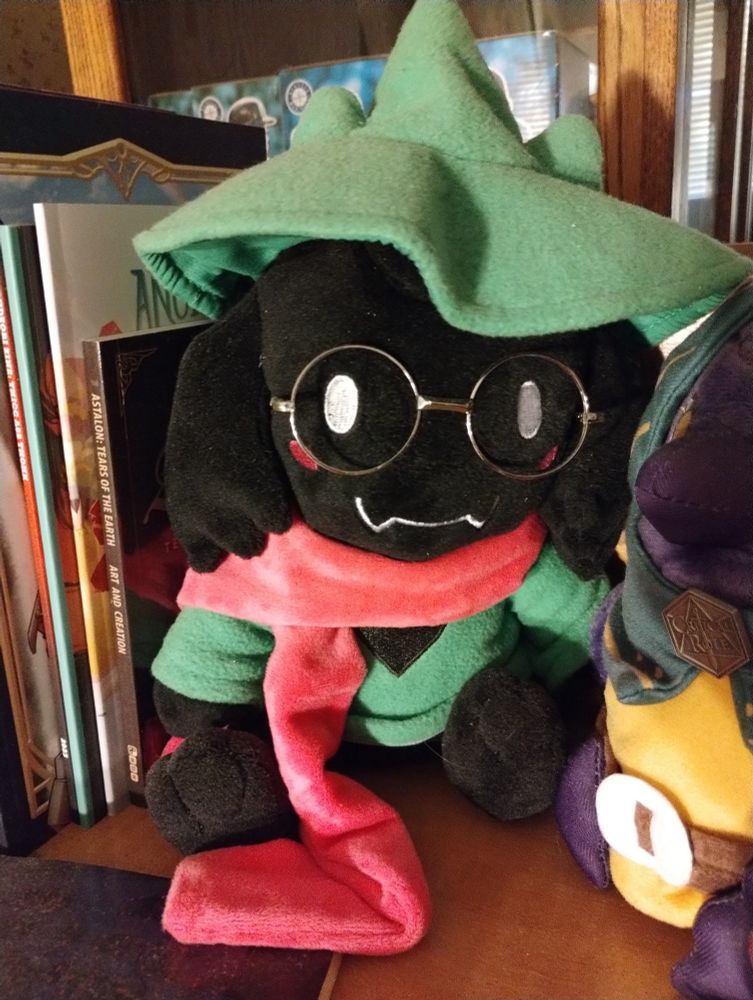 a Hat Ralsei plush, sitting on a shelf next to some friends. 
