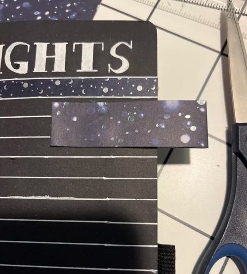 The corner of a black paper bullet journal page. A magnetized tab bookmark made of starry paper is clipped to it.