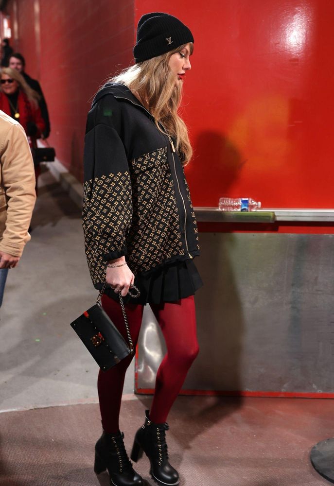 Taylor Swift arriving to Arrowhead Stadium wearing black heel combat boots, red opaque tights, a black pleated mini skirt, and a black LV logo hoodie and black beanie with LV logo printed on the side. Her hair is down and in loose waves, and from her side profile we see she is wearing red lipstick and black winged eyeliner. She's carrying a small black rectangular leather purse in her hand