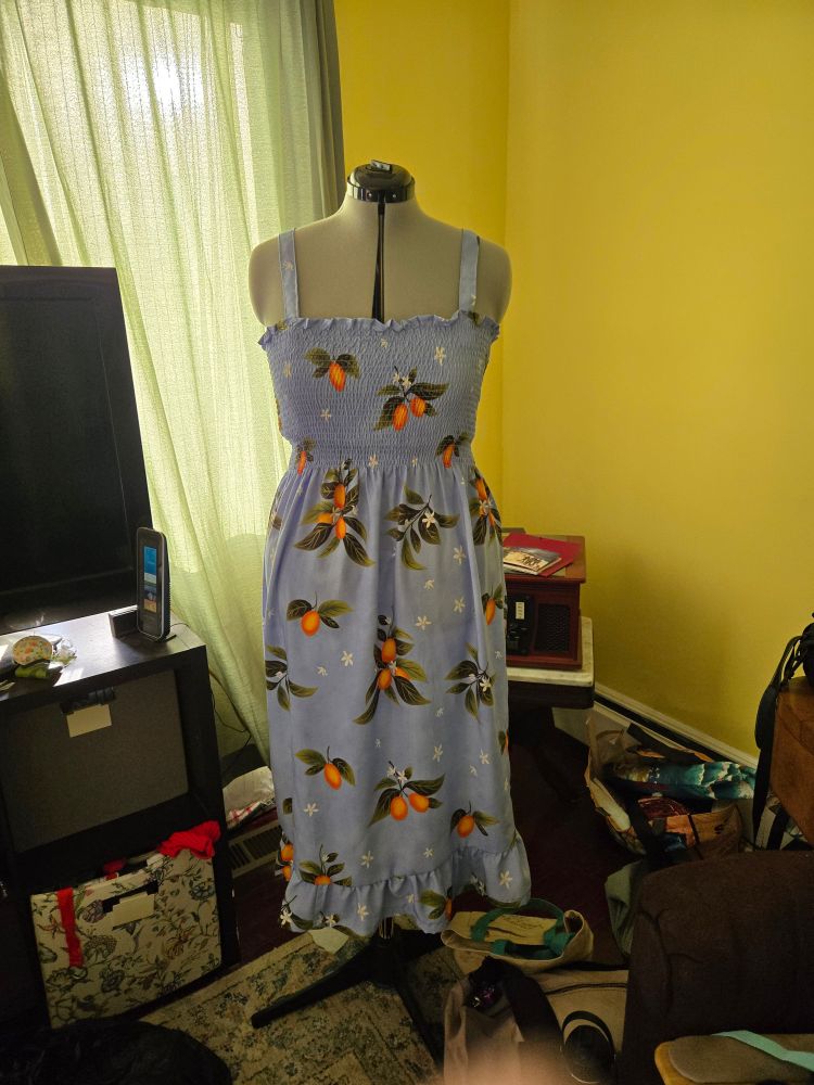 A sundress in sky blue with oranges and springs of orange blossoms printed on the fabric on a dressform.