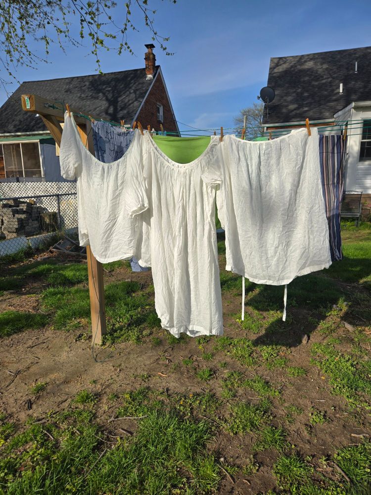 Two different white linen chemises and one linen underpetticoat hanging on a laundry line in the yard.