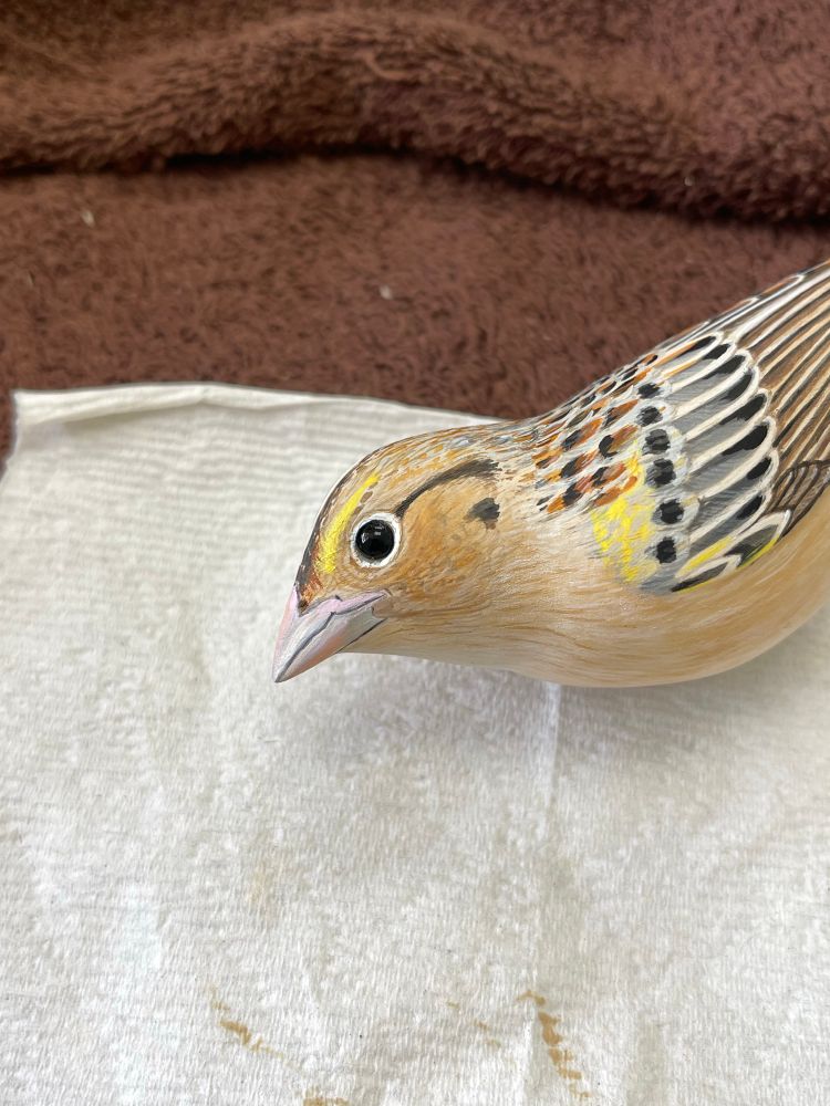 Small bird painted with a tans, yellows, chestnut, grays, and black. With a pinkish beak and a black glass eye. 