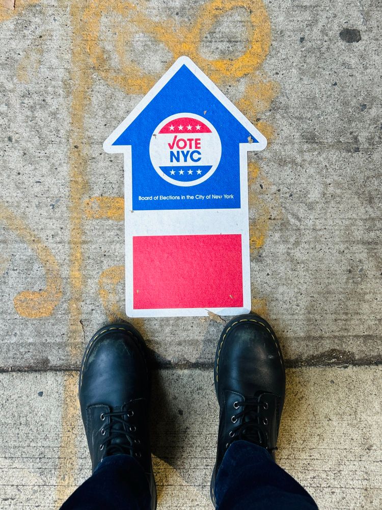 boots walking toward a red white and blue arrow sticker on the sidewalk, printed w the words: 
NYC
VOTE NYC
****
Board of Elections in the City of Now York