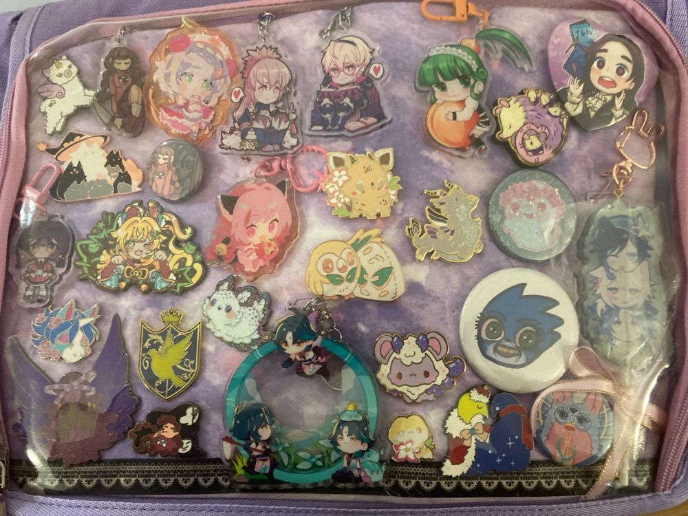 In a purple itabag, on a marbled purple and pink backing, is a wide array of pins and keychains.
There’s items based off series including Genshin Impact, Vocasynth/Vocaloid, Hatoful Boyfriend, Pokemon, Yume Nikki, Fire Emblem, Animal Crossing, Dream Daddy, a jesus furby, a cursed original movie design sonic, and some original cute and pretty pins.