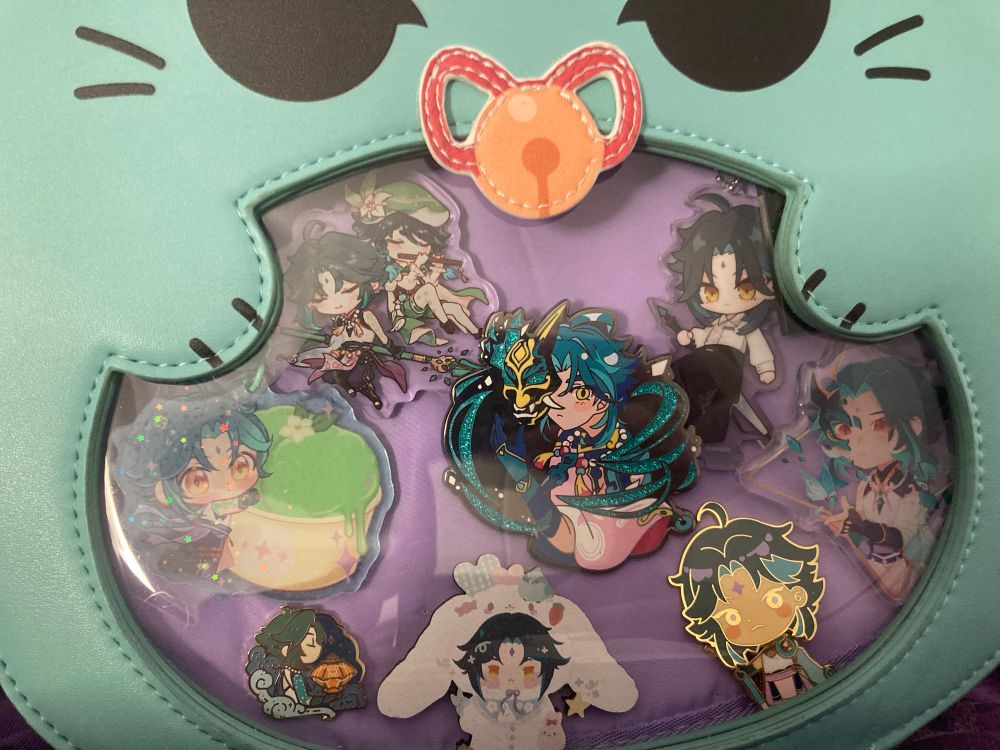 In a green, cat Xiao-shaped itabag, on a purple backing, are a bunch of Xiao pins and keychains.